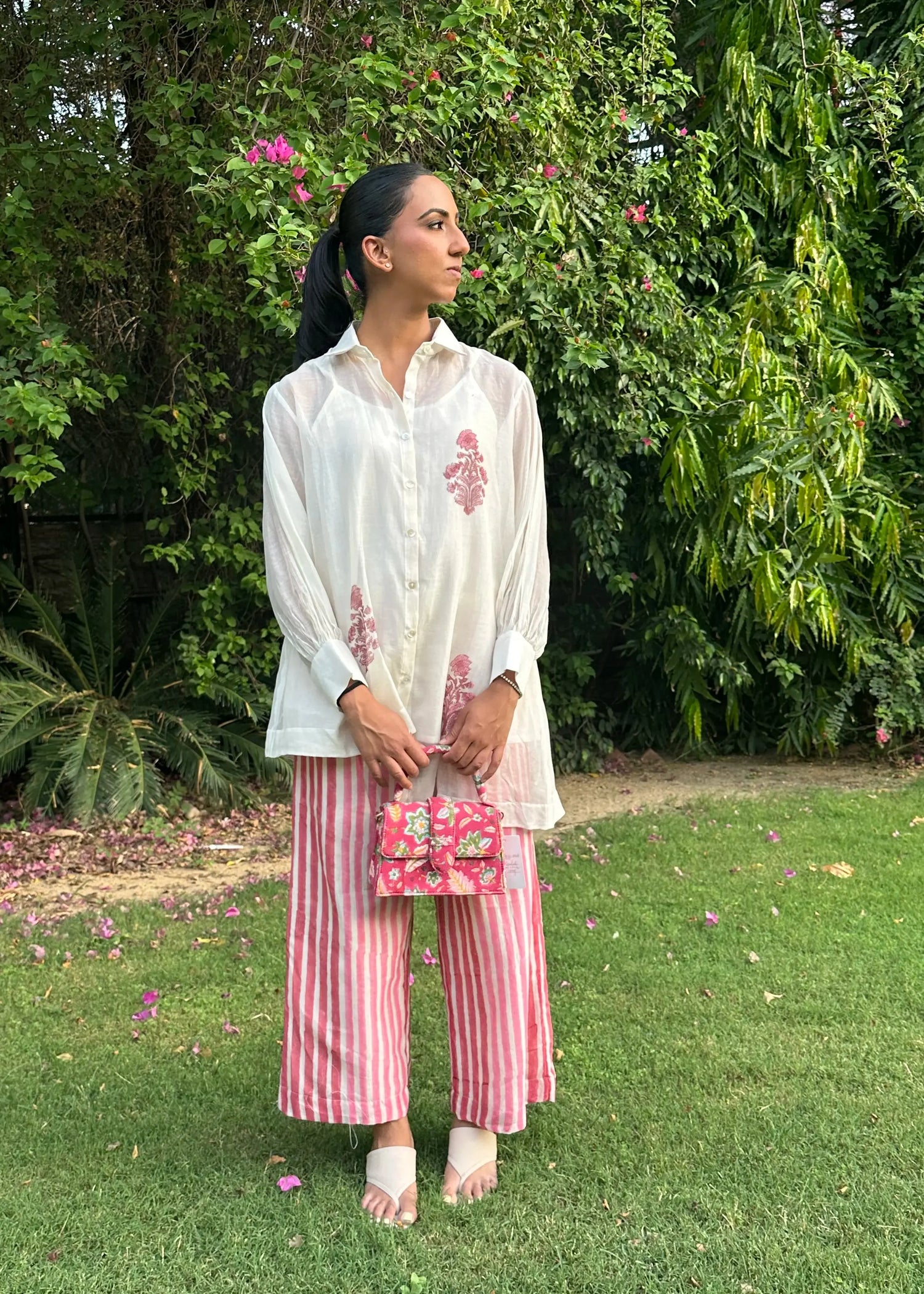 A woman stands on a grassy lawn in front of dense green foliage, wearing a white embroidered long-sleeve top, pink and white striped wide-leg pants, and holding a small pink floral handbag.