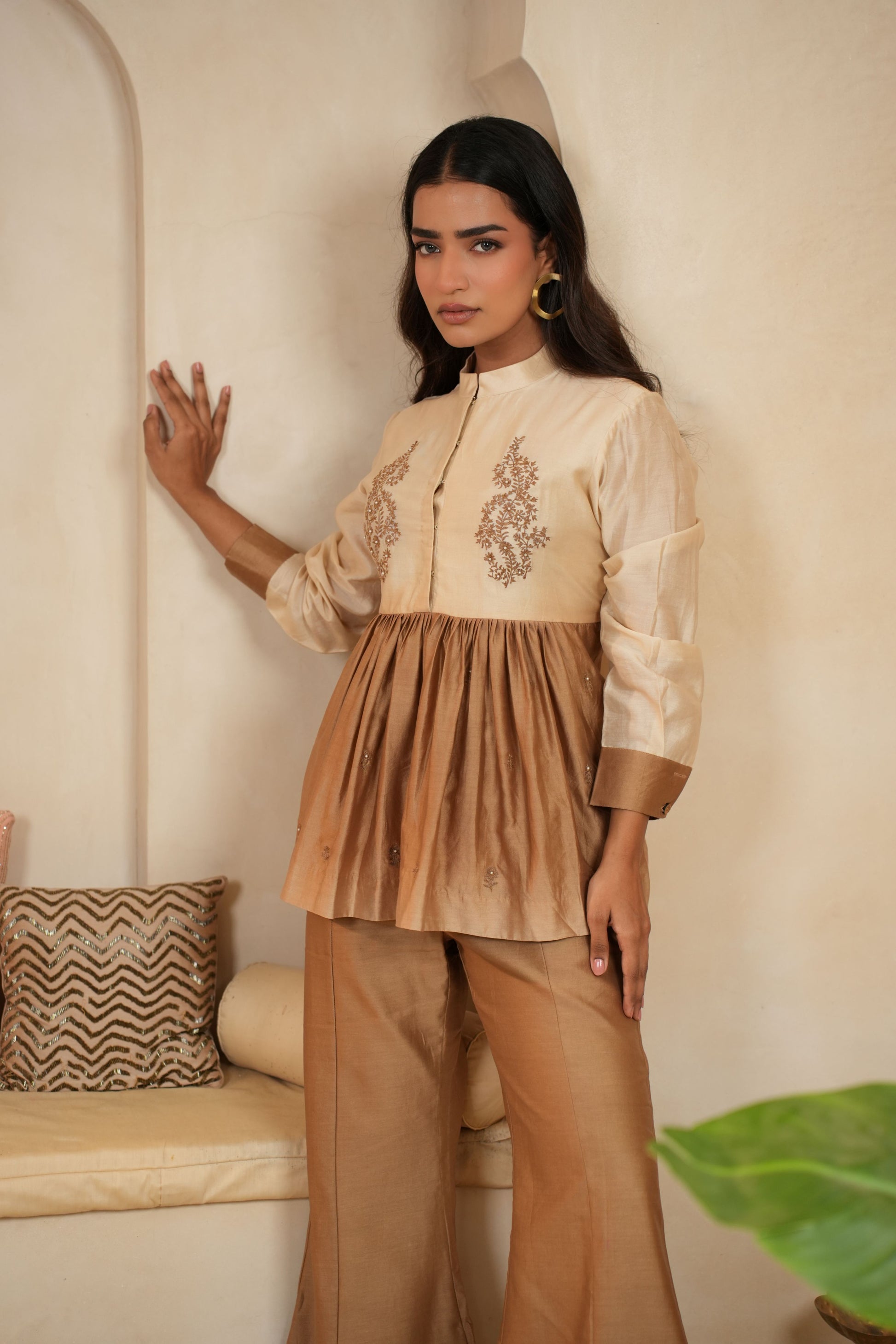 Woman wearing a beige embroidered top and pants in a room with a decorative pillow and plant.