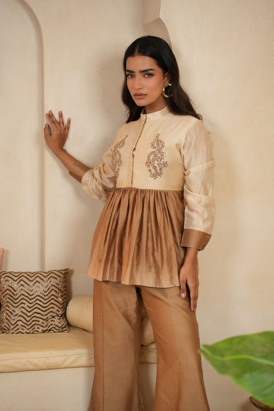 Woman wearing a beige embroidered top and pants in a room with a decorative pillow and plant.