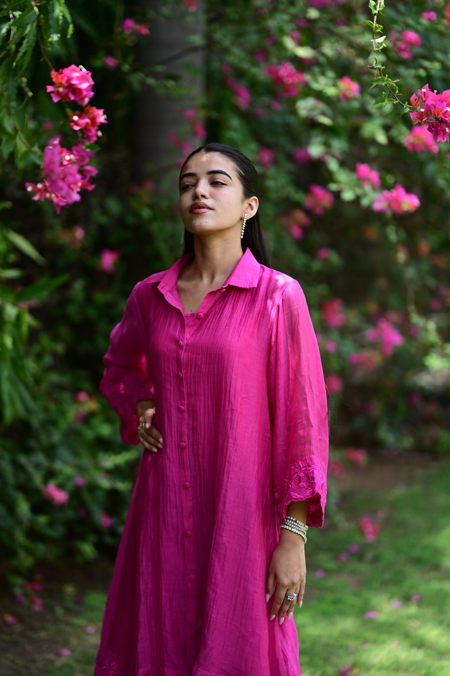A woman wearing a vibrant magenta pink long-sleeve button-up shirt dress, standing in a lush garden with pink flowers and green foliage.