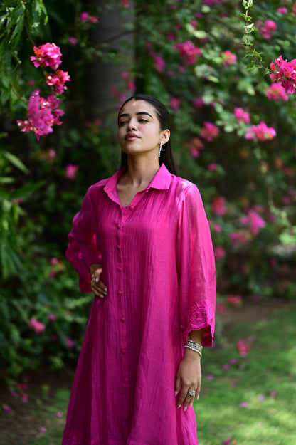 A woman wearing a vibrant magenta pink long-sleeve button-up shirt dress, standing in a lush garden with pink flowers and green foliage.