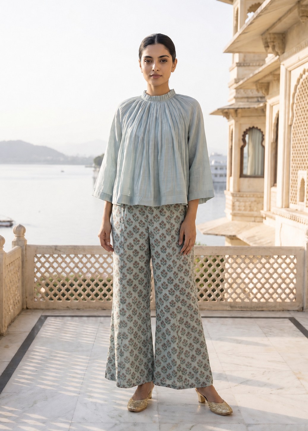 Woman in light blue top and patterned pants standing on a balcony with a scenic view.