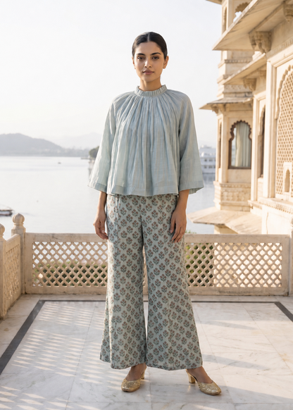Woman in light blue top and patterned pants standing on a balcony with a scenic view.