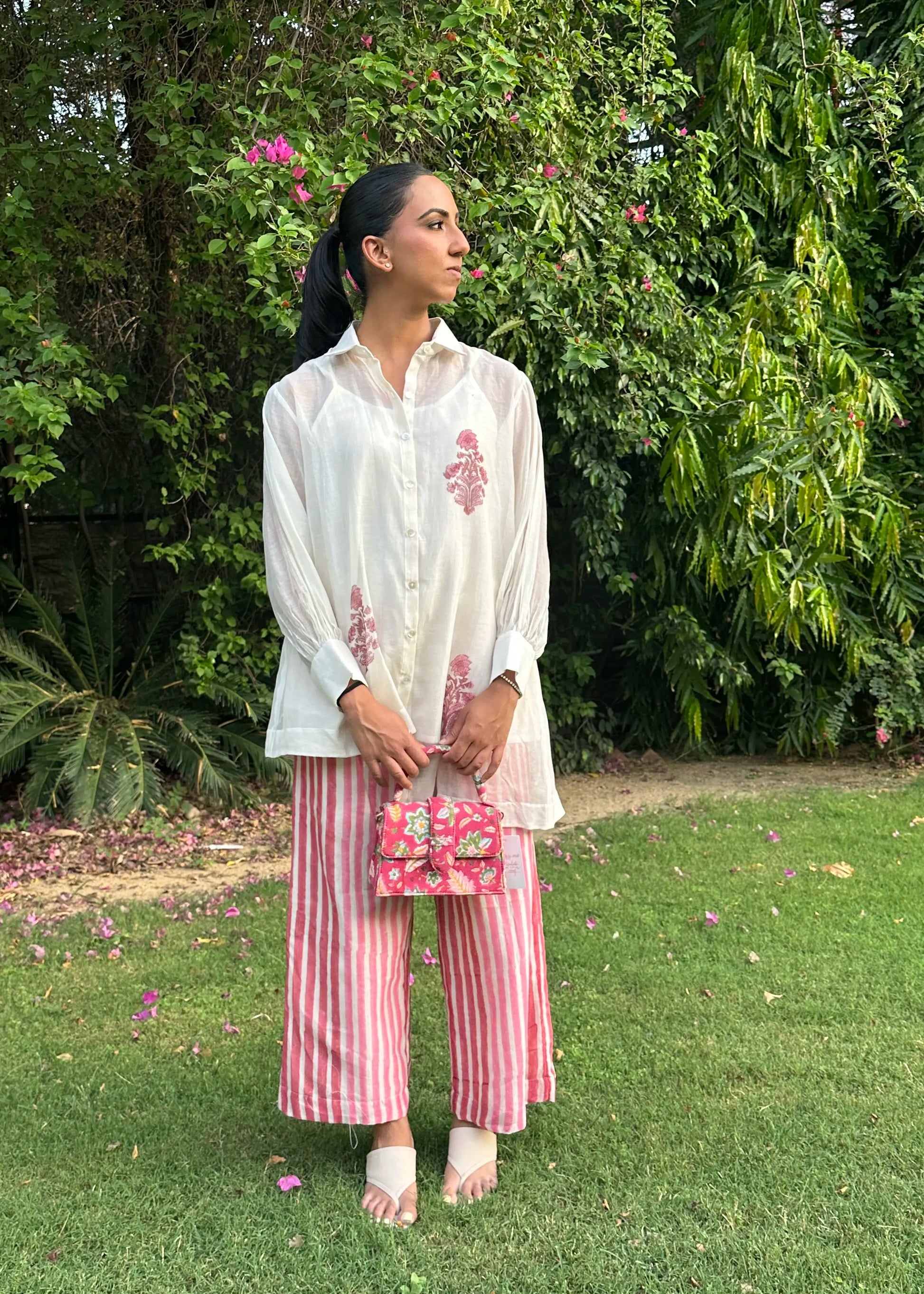 A woman stands on a grassy lawn in front of dense green foliage, wearing a white embroidered long-sleeve top, pink and white striped wide-leg pants, and holding a small pink floral handbag.