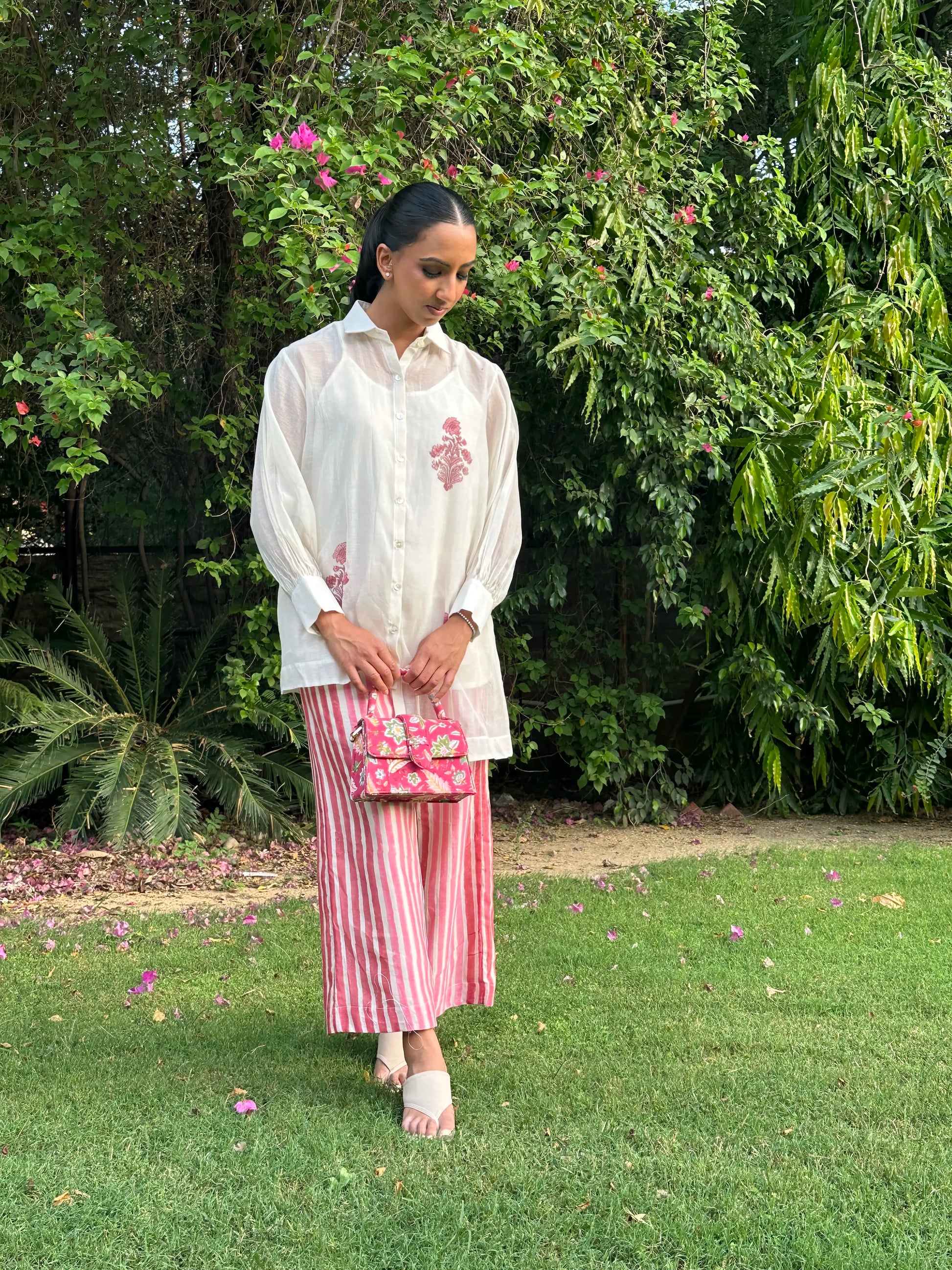 A woman stands on a grassy lawn in front of lush green foliage, wearing a white embroidered long-sleeve shirt, pink and white vertically striped wide-leg pants, and holding a pink patterned handbag.