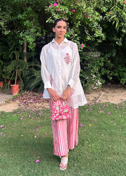 A woman stands on a grassy lawn in front of dense green foliage and flowering plants, wearing a loose white long-sleeve top and pink vertically striped pants, holding a pink floral handbag.