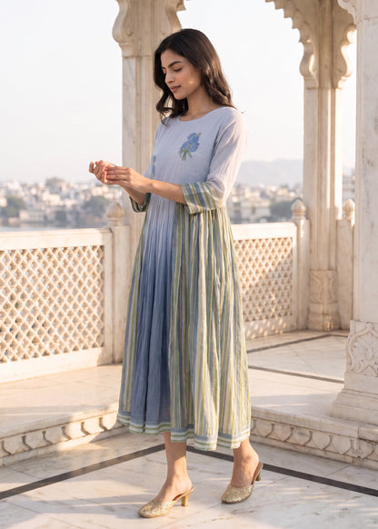 A woman models a light blue and green striped maxi dress with a blue floral embroidery on the chest, standing on a marble terrace with a scenic background.