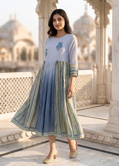 A woman models a light blue and green striped maxi dress with floral embroidery on the chest, standing on a marble terrace with a domed palace in the background.