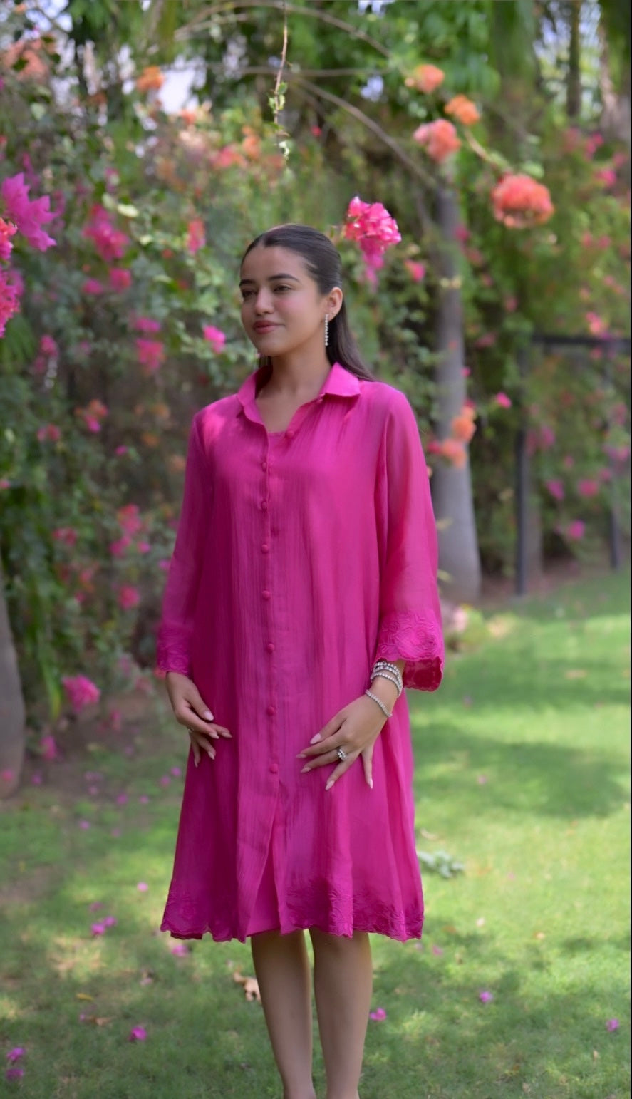 A woman wearing a bright pink chanderi shirt dress stands in a garden with green grass and pink flowers in the background.