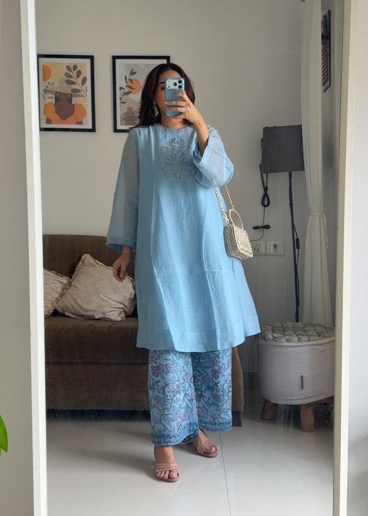 A woman takes a mirror selfie in a living room, wearing a light blue long-sleeve tunic top with matching patterned wide-leg pants. She is holding a phone to take the photo, with a brown sofa and framed wall art in the background.